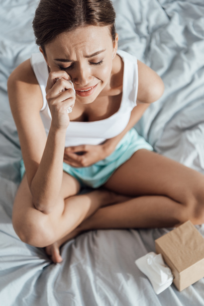 korkea kulma näkymä stressaantunut nuori nainen koskettaa vatsa ja itkee sängyssä
 - Valokuva, kuva
