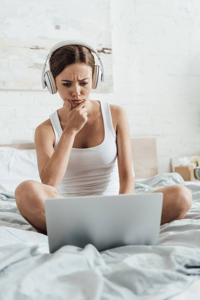 pensive young woman listening music in headphones and using laptop on bed - Photo, Image