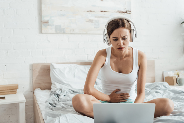 stressed woman in headphones touching belly and using laptop on bed - Zdjęcie, obraz