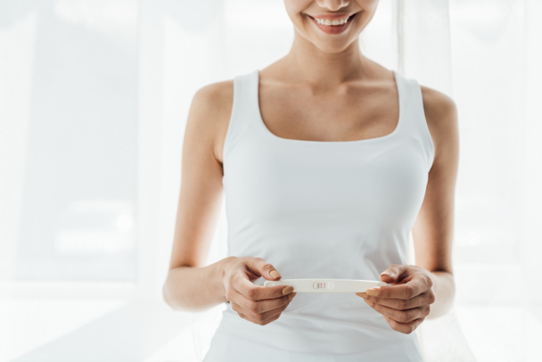 обрезанный вид улыбающейся молодой женщины, проводящей тест на беременность дома
 - Фото, изображение