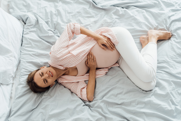 overhead view of pregnant woman lying in bed with grimace and touching belly - Foto, afbeelding