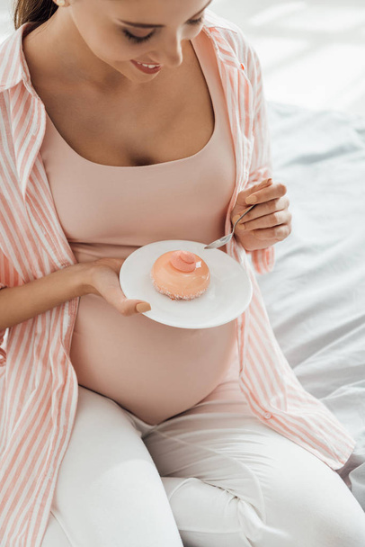 smiling pregnant woman sitting on bed and holding saucer with cupcake - Valokuva, kuva