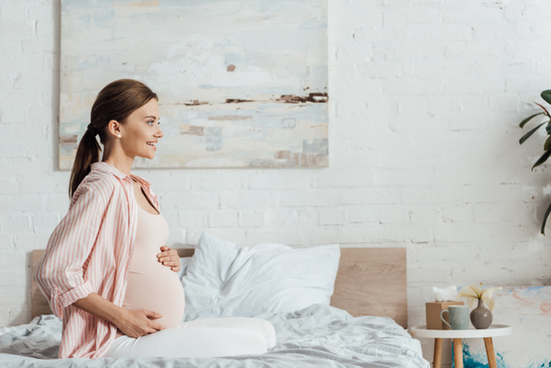 side view of happy pregnant woman sitting on bed and touching belly - Valokuva, kuva