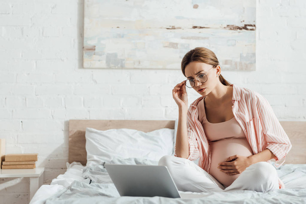 zwangere vrouw in glazen zittend op bed en met behulp van laptop - Foto, afbeelding