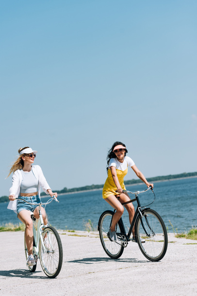 blonde and brunette girls riding bikes near river in summer - Foto, afbeelding