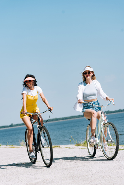 smiling blonde and brunette girls riding bikes near river in summer - Fotó, kép