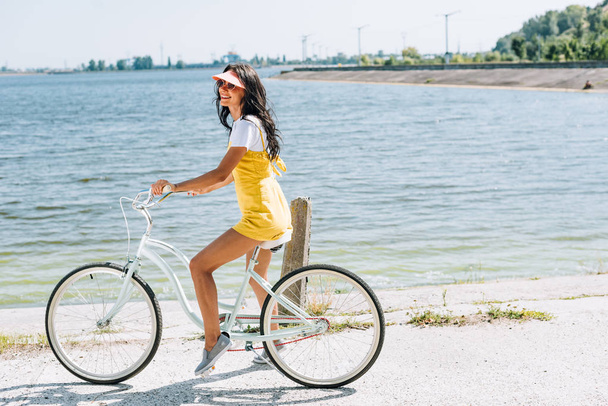 side view of happy brunette beautiful girl riding bicycle near river in summer - Foto, Imagen
