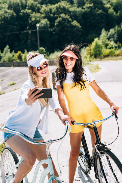 blonde and brunette girls riding bikes and taking selfie in summer - Фото, изображение