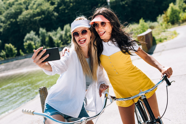 selektiver Fokus von blonden und brünetten Mädchen, die Zunge zeigen, während sie Selfies auf Fahrrädern machen - Foto, Bild