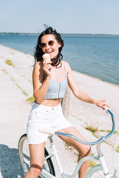 brunette girl eating ice cream and riding bike near river in summer - Foto, Imagen