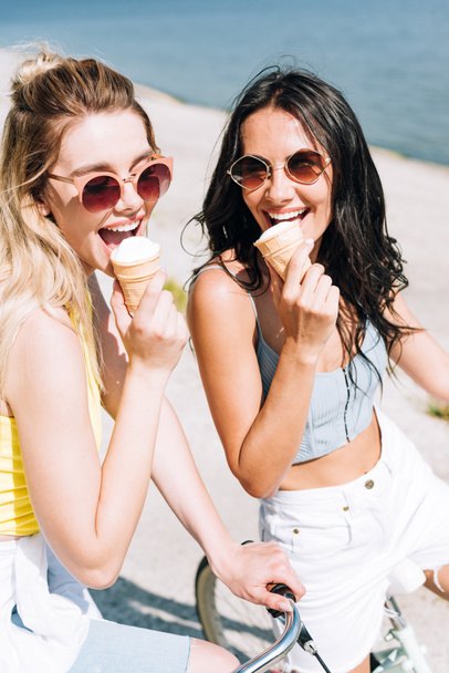 happy blonde and brunette girls riding bikes and eating ice cream - Фото, зображення