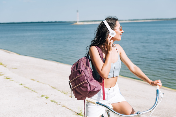 happy girl with backpack biking and listening music in headphones near river in summer - Foto, Imagem