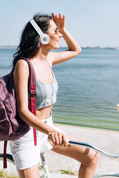 happy brunette girl with backpack biking and listening music in headphones near river - Photo, Image