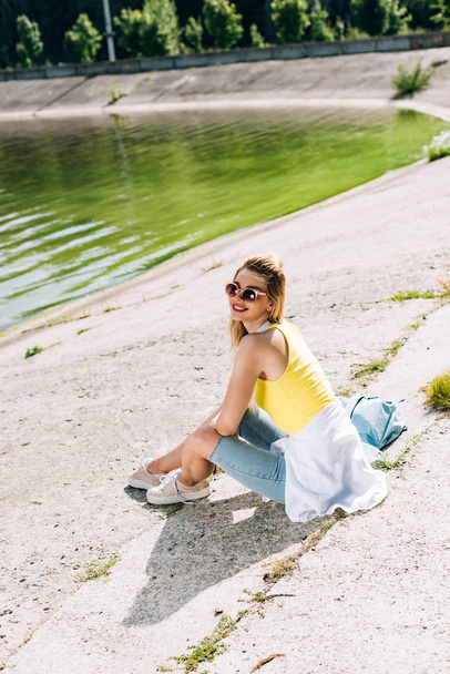 happy blonde girl sitting near river in summer - Photo, Image