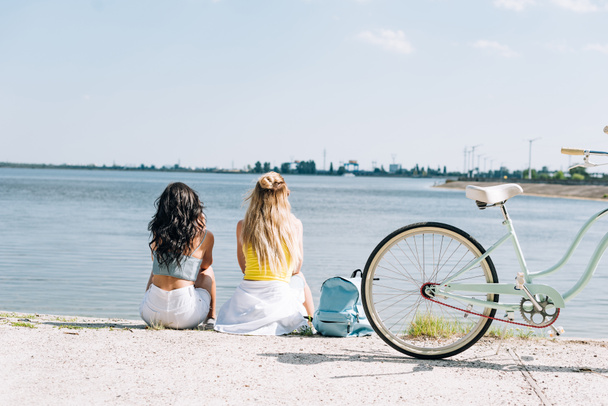 back view of blonde and brunette friends sitting near bike and river in summer - Фото, изображение