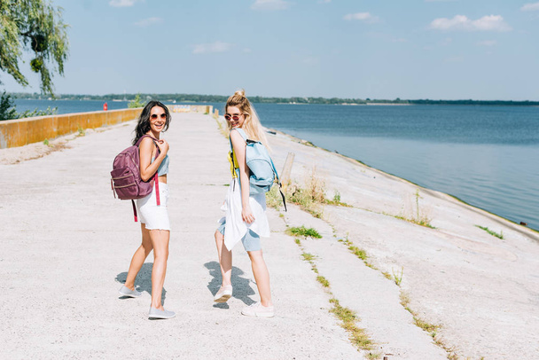 happy blonde and brunette girls walking with backpacks near river in summer - Photo, image