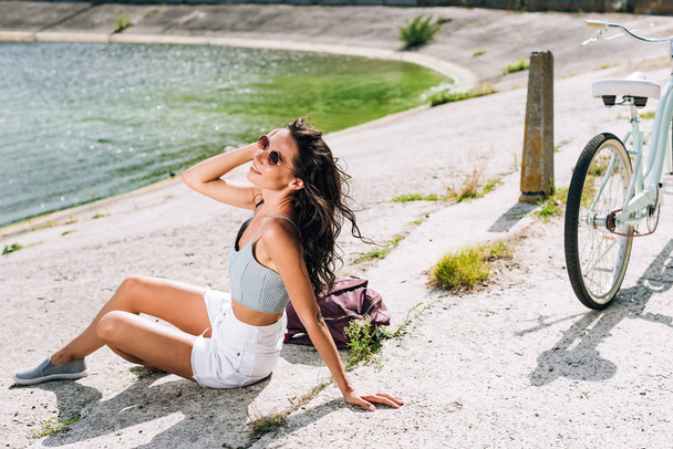 brunette girl with bike sitting near river in summer - Zdjęcie, obraz