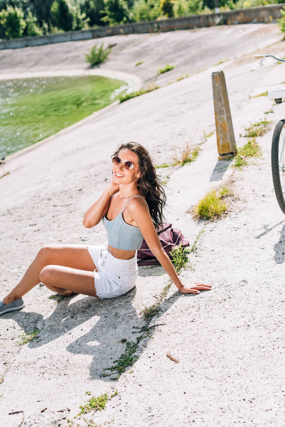 beautiful brunette girl with bike sitting near river in summer - Фото, изображение