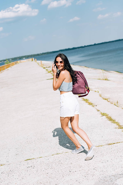 szczęśliwy brunetka dziewczyna w okulary przeciwsłoneczne chodzenie z plecak blisko rzeka w lato - Zdjęcie, obraz