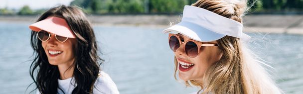 panoramic shot of happy blonde and brunette girls in sunglasses near river - Фото, изображение