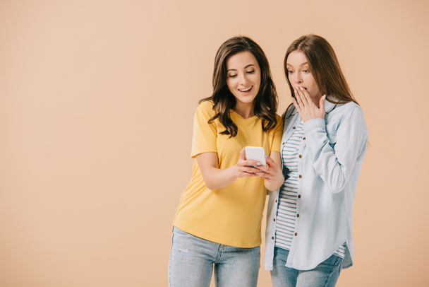 attractive woman looking at smartphone with her shocked friend isolated on beige - Photo, Image