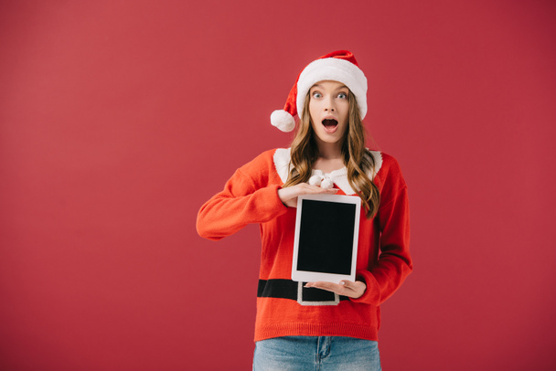 attractive woman in santa hat and sweater holding digital tablet isolated on red - 写真・画像
