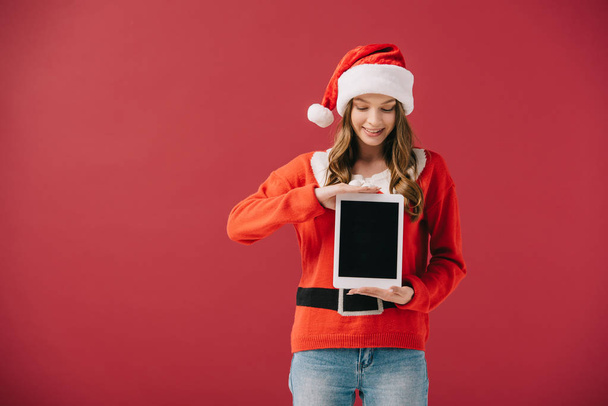 attractive woman in santa hat and sweater holding digital tablet isolated on red - 写真・画像