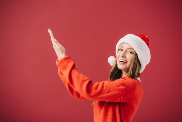 ελκυστική γυναίκα σε Σάντα καπέλο και πουλόβερ που δείχνουν με το χέρι απομονωμένο στο κόκκινο  - Φωτογραφία, εικόνα