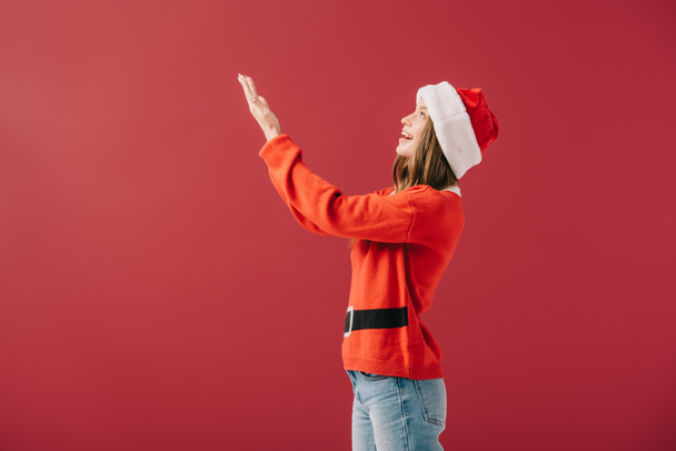 side view of attractive woman in santa hat and sweater pointing with hand isolated on red  - Фото, изображение