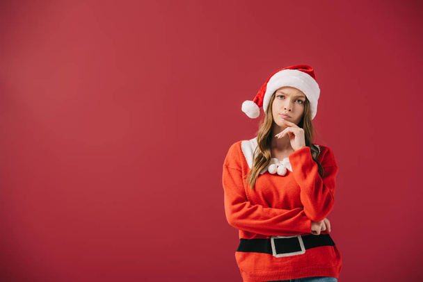 attractive and pensive woman in santa hat and sweater looking at camera isolated on red  - Foto, Imagem
