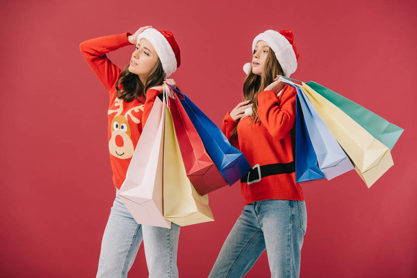 attractive women in sweaters and santa hats holding shopping bags and looking away isolated on red  - Фото, изображение
