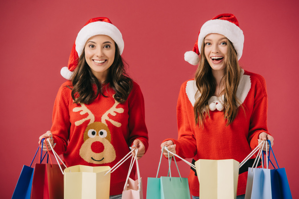 des femmes attirantes et souriantes dans des pulls et des chapeaux de Père Noël tenant des sacs à provisions isolés sur rouge
  - Photo, image