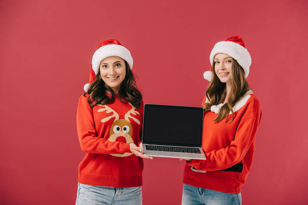 attractive and smiling women in sweaters and santa holding laptop isolated on red  - 写真・画像