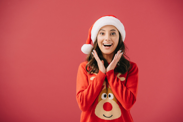 atraktivní a usmívající se ženy na svetry a na Santa klobouky, které se dívají na kameru izolovaně na červenou  - Fotografie, Obrázek