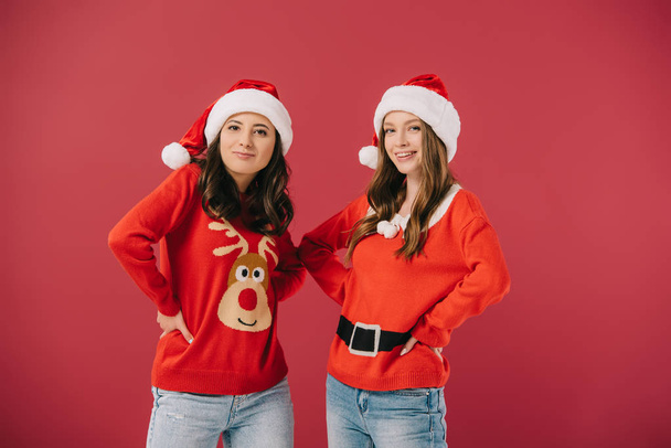 attractive and smiling women in sweaters and santa hats with hands on hips looking at camera isolated on red  - Foto, immagini