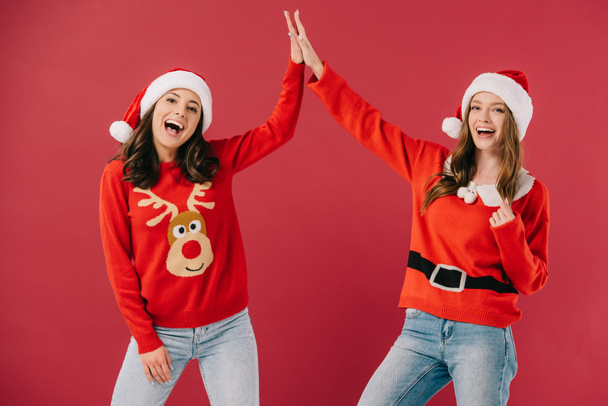 attractive and smiling women in sweaters and santa hats giving high five isolated on red  - Фото, изображение