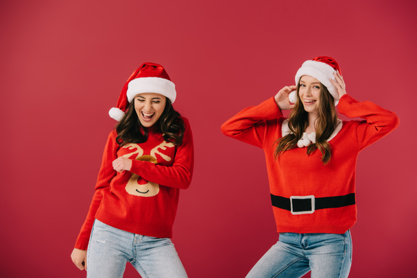 attractive and smiling women in sweaters and santa hats looking at camera isolated on red  - Foto, afbeelding