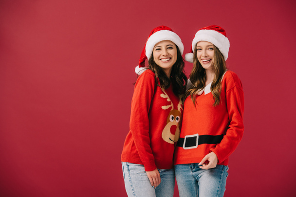 aantrekkelijke en lachende vrouwen in truien en kerstman hoeden kijken naar camera geïsoleerd op rood  - Foto, afbeelding