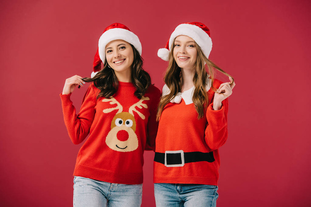 atrakcyjne i uśmiechnięte kobiety w swetry i Santa kapelusze patrząc na aparat na białym tle na czerwono  - Zdjęcie, obraz