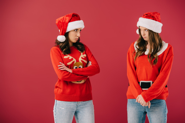 attractive and offended women in sweaters and santa hats looking at each other isolated on red  - Photo, Image