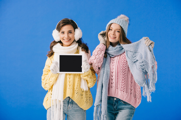 mulheres atraentes e sorridentes em suéteres e lenços segurando tablet digital e olhando para a câmera isolada em azul
 - Foto, Imagem