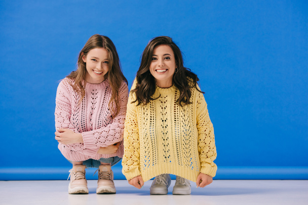 attractive and smiling women in sweaters looking at camera on blue background  - Zdjęcie, obraz