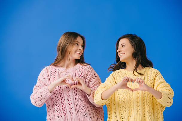 attractive and smiling women in sweaters showing heart gesture isolated on blue  - Photo, image