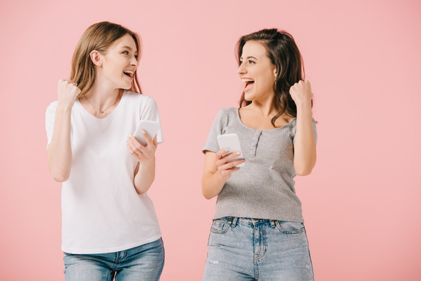 mujeres atractivas y sonrientes en camisetas con teléfonos inteligentes y mostrando sí gesto aislado en rosa
 - Foto, imagen
