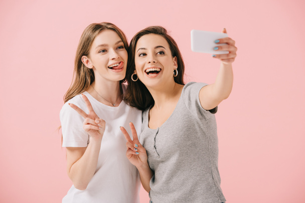 mujeres atractivas y sonrientes en camisetas tomando selfie y mostrando signos de paz aislados en rosa
 - Foto, imagen