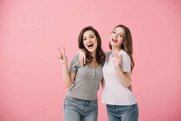 mujeres atractivas y sonrientes en camisetas mirando a la cámara y mostrando un signo de paz aislado en rosa
 - Foto, Imagen