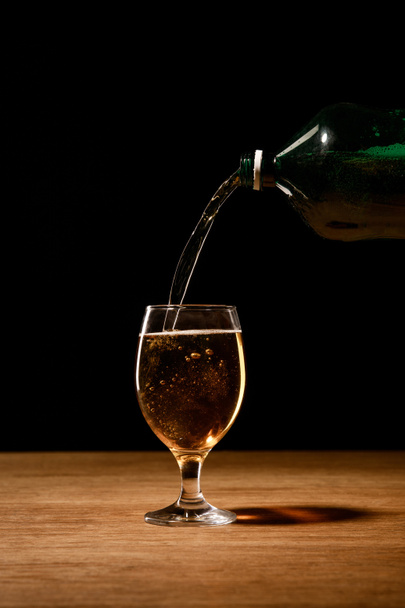 beer pouring from bottle in glass on wooden table isolated on black - Fotografie, Obrázek