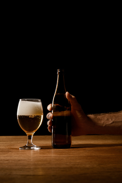 gedeeltelijke weergave van man Holding fles in de buurt van glas met bier en schuim op houten tafel geïsoleerd op zwart - Foto, afbeelding