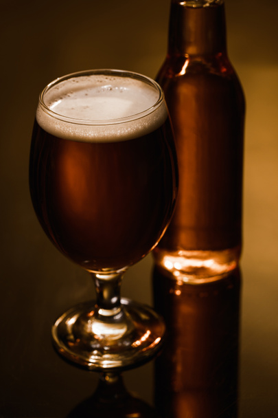 selektiver Fokus des Bieres mit Schaum im Glas in der Nähe der Flasche auf dunklem Hintergrund mit Beleuchtung - Foto, Bild