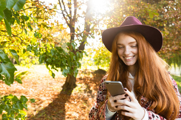 Smiling cutie young student redhead girl in autumn park using mobile phone. - Фото, зображення